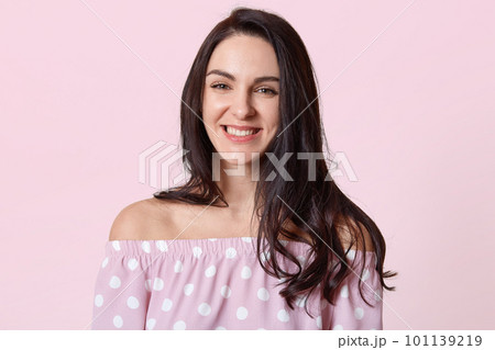 Portrait of beautiful woman with dark hair, smiles broadly, being in good mood, dressed in polka dot dress, poses against rosy studio wall, has healthy skin, minimal make up. Emotions concept Portrait of beautiful woman with dark hair, smiles broadly, being in good mood, dressed in polka dot dress, poses against rosy studio wall, has healthy skin, minimal make up. Emotions concept 101139219