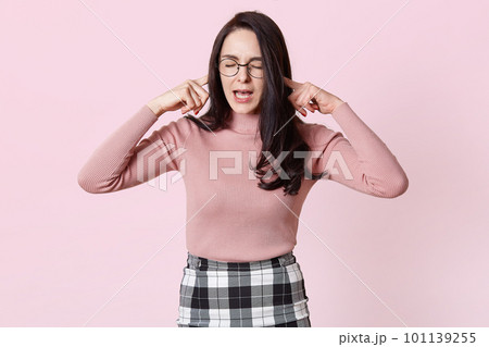 Beautiful grimacing young woman in pink sweater, her ears with fingers and she keeps eyes closed. Charming female with long dark hair wears round glasses, do not want hear any word. People concept. 101139255