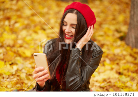 Emotional portrait of happy and relaxed beautiful young woman with dark hair, dresses beret and jacket, looks smiling at phone while sitting on fallen yellow leaves during walk in park. Autumn colors. 101139271