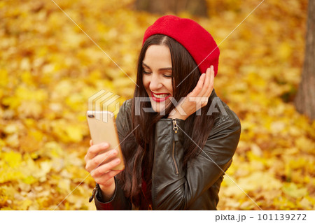 Close up outdoor portrait of woman, lady looks happy and taking self picture via smartphone in autumn park, young model posing on ground covering golden leaves. Technology and happiness concept. 101139272