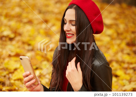 Indoor shot of woman taking selfie via mobile phone in fall. Autumn girl having smartphone video conversation. Portrait of Caucasian female in forest in fall colors, lady wearing red beret and jacket. 101139273