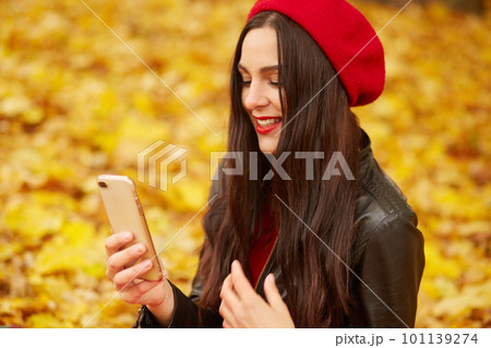 Close up portrait of female wearing red beret and leather jacket, model sitting on ground outside. Young attarctive woman spending time in autumn park, looking at telephone screen with gentle smile. 101139274