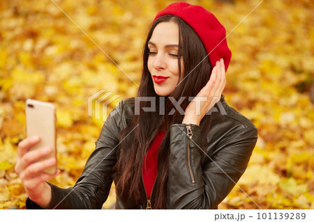Portrait of cheerful young woman surrounded with autumn leaves, posing in fall park full of foliage, making selfie via her smartphone, dresses red beret and black leather jacket. People concept. Portrait of cheerful young woman surrounded with autumn leaves, posing in fall park full of foliage, making selfie via her smartphone, dresses red beret and black leather jacket. People concept. 101139289