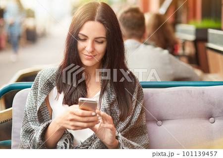 Online internet connection concept. Beautiful young brunette lady uses mobile phone for searching internet, wrapped in coverlet, spends free time in coffee shop, recieves message from boyfriend Online internet connection concept. Beautiful young brunette lady uses mobile phone for searching internet, wrapped in coverlet, spends free time in coffee shop, recieves message from boyfriend 101139457