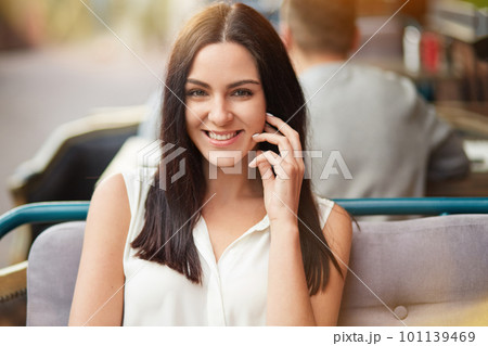 People, beauty and happiness concept. Attractive brunette female in white blouse, has delighted expression, healthy skin, sits against blurred outdoor cafe background, smiles broadly at camera People, beauty and happiness concept. Attractive brunette female in white blouse, has delighted expression, healthy skin, sits against blurred outdoor cafe background, smiles broadly at camera 101139469