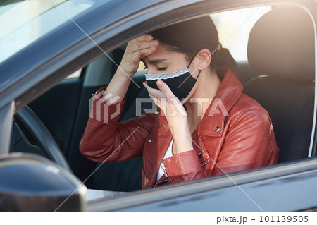 Picture of infected sick young female closing eyes, coughing, covering mouth, having fever, putting one hand on forehead, driving car, being ill with coronavirus. People and covid 19 concept. 101139505
