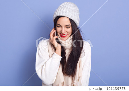 Happy young woman talking to her friend via smart phone isolated over blue background, lady with red lips dresses white warm clothes, brunette girl having joyful facial expression. People concept. 101139773