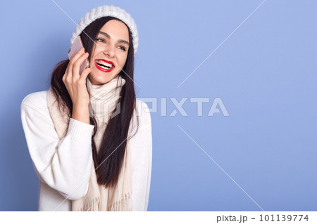 Close up portrait of beautiful happy woman dresses warm clothes, smiling and using cellphone, talk to his boyfriend, poses isolated over blue background. Copy space for advertisment or promotion. Close up portrait of beautiful happy woman dresses warm clothes, smiling and using cellphone, talk to his boyfriend, poses isolated over blue background. Copy space for advertisment or promotion. 101139774