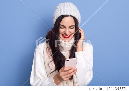 Portrait of nice attractive satisfied cheerful girl holding in hands and using cell phone, lady stands isolated over bright blue background, female wearing white pullover, scarf and cap. 101139775