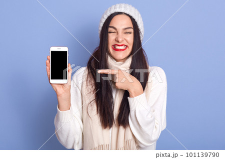 Horizontal shot of woman showing smart phone with blank screen and pointing at It, posing isolated over blue background, winsome lady wearing white warm sweater and cap. Copy space for advertisment. Horizontal shot of woman showing smart phone with blank screen and pointing at It, posing isolated over blue background, winsome lady wearing white warm sweater and cap. Copy space for advertisment. 101139790