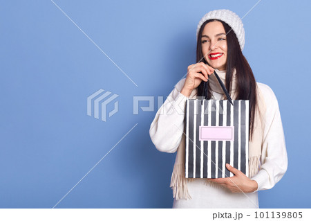 Happy dark haired girl with bright red lips looking away and smiling, swing striped shopping bag, wearing warm clothing, copy space for advertisement or promo, posing against blue wall. 101139805