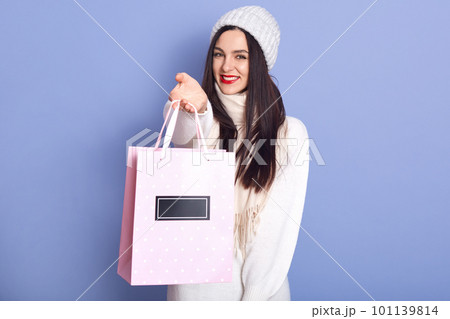 Indoor studio shot of cheerful beautiful female wearing white sweater, hat and scarf, holding paperbag with Christmas present, smiling sincerely, standing isolated over lilac background. Winter time. 101139814