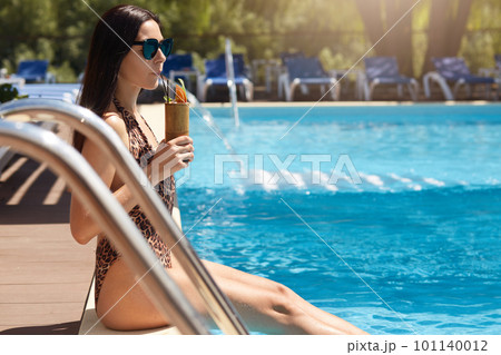 Stylish lady resting at resort, sitting near swimming pool with legs in water on vacation, attractive woman dressed fashionable swimsuit and black sunglasses, looking at distance. Recreation concept. 101140012