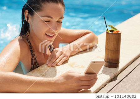 Portrait of attentive beautiful female being in clean swimming pool, holding her device in one hand, looking at smartphone screen carefully, checking social networking sites, having cocktail nearby. Portrait of attentive beautiful female being in clean swimming pool, holding her device in one hand, looking at smartphone screen carefully, checking social networking sites, having cocktail nearby. 101140082