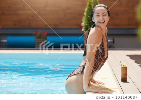 Side view of young woman relaxing in swimming pool. Beautiful Caucasian female with wet hair in swimsuit with leopard print enjoys sunny days at resort. Outdoor hotel pool and summer vacation concept 101140087
