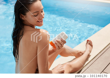 Attractive woman putting solar cream or sunblock on her armor shoulder, smiling lady dresses bikini by pool. Beautiful young woman under sun on sunny summer day, relaxing at luxury spa resort. Attractive woman putting solar cream or sunblock on her armor shoulder, smiling lady dresses bikini by pool. Beautiful young woman under sun on sunny summer day, relaxing at luxury spa resort. 101140170