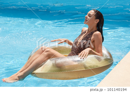 Side view og slim tanned woman in swimming suit with leopard print, looks while lyingon inflatable ring in swimming pool, spending hot summer days at luxury resort. Recreation and vacation concept. 101140194
