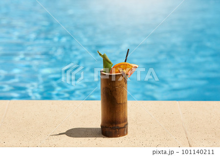 Exotic cocktail in wooden glass with lemon and drinking tube standing on poolside with blue pool's water on background, tasty fresh beverage on resort. 101140211