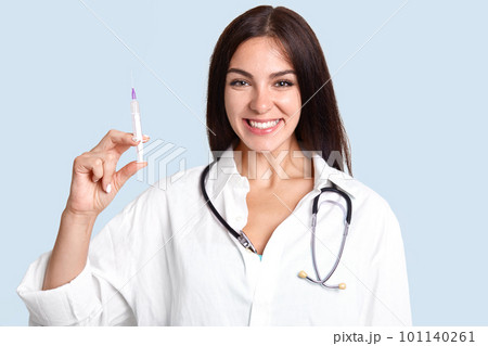 Cheerful brunette female doctor with stethoscope and syringe going to give medical treatment to patient, has friendly look, isolated over blue background. Woman general practitioner in hospital Cheerful brunette female doctor with stethoscope and syringe going to give medical treatment to patient, has friendly look, isolated over blue background. Woman general practitioner in hospital 101140261