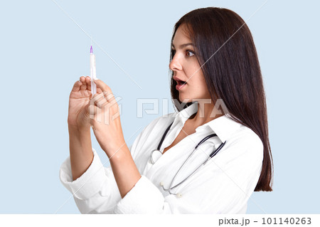 Sideways shot of female physician holds syringe, gong to make vaccination, looks at remedy with shocked expression, poses against light blue background, being professional specialist in medical sphere Sideways shot of female physician holds syringe, gong to make vaccination, looks at remedy with shocked expression, poses against light blue background, being professional specialist in medical sphere 101140263