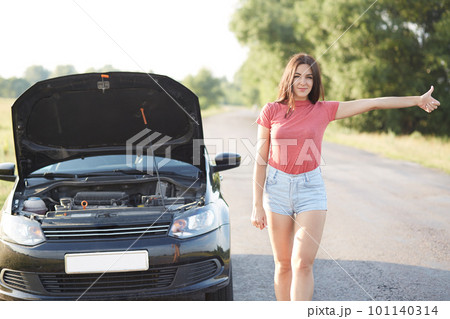 People and problems with transport. Attractive unexperienced female driver makes stop gesture on road, stands near brocken car, feels frustrated as doesn`t know how repair it, has trouble with engine 101140314