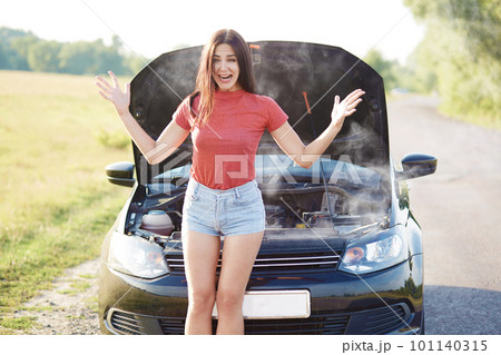 Woman in despair raises hands, stand near brocken automobile, smoke from hood, faces technical problem, wears casual t shirt and jean shorts, has slender legs, stops on road. Transport concept 101140315