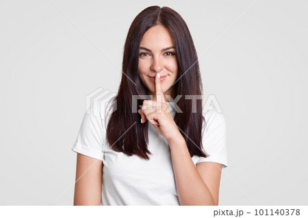 Attractive woman with long dark hair has secret, keeps fore finger over mouth, shows shush gesture, has satisfied facial expression, dressed in casual outfit, isolated over white background. Attractive woman with long dark hair has secret, keeps fore finger over mouth, shows shush gesture, has satisfied facial expression, dressed in casual outfit, isolated over white background. 101140378