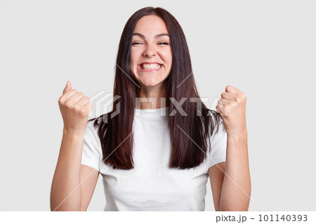Successful woman clenches fists with happiness, has broad smile, squints face, dressed casual t shirt, isolated over white background. Carefree brunette girl rejoices success. Victory, determination Successful woman clenches fists with happiness, has broad smile, squints face, dressed casual t shirt, isolated over white background. Carefree brunette girl rejoices success. Victory, determination 101140393