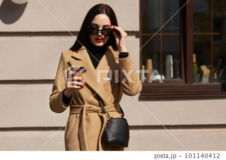 Outdoor shot of attractive young woman wearing coat in sunny city street and drinking take away coffee in paper cup, keeps hand on her sunglasses, going to office after lunch breack. People concept. Outdoor shot of attractive young woman wearing coat in sunny city street and drinking take away coffee in paper cup, keeps hand on her sunglasses, going to office after lunch breack. People concept. 101140412