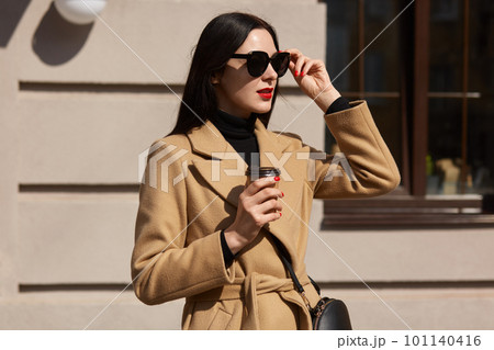 Outdoor fashion photo of young beautiful isolated over street building background, elegant lady lifting her sunglasses and looking away, enjoys drinking hot takeaway beverage while walking alone. 101140416