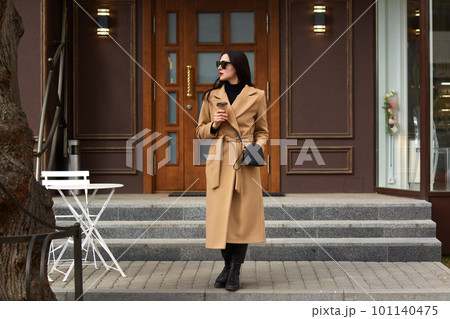 Beautiful young brunettee woman in modern beige coat, holding disposable takeaway cup and standing against urban city building, looks thoughtfuly aside, has dating, waiting for her boyfriend. Beautiful young brunettee woman in modern beige coat, holding disposable takeaway cup and standing against urban city building, looks thoughtfuly aside, has dating, waiting for her boyfriend. 101140475