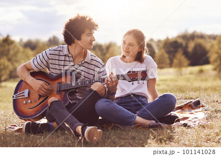 Beautiful female looks with love and happiness at her boyfriend who plays guitar and sings romantic songs to lover, have unforgettable date outdoor, enjoy togetherness. People, love, relations 101141028