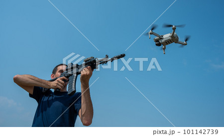 A man aims to shoot a rifle at a flying drone against a blue sky.  101142739