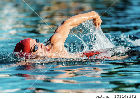 Male swimmer swimming crawl freestyle in blue water outdoor pool. Portrait of an athletic young male triathlete swimming crawl wearing a red cap and swim goggles. Triathlete training for triathlon Male swimmer swimming crawl freestyle in blue water outdoor pool. Portrait of an athletic young male triathlete swimming crawl wearing a red cap and swim goggles. Triathlete training for triathlon 101143382