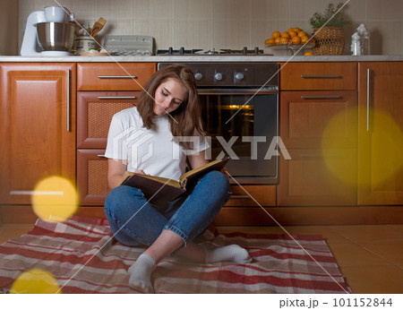 Horizontal shot of a happy woman sitting on the floor in the kitchen with an open notepad waiting for a delicious pie baking in the oven Horizontal shot of a happy woman sitting on the floor in the kitchen with an open notepad waiting for a delicious pie baking in the oven 101152844