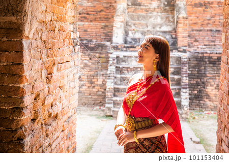Young woman dressed in traditional red Thai dress and golden ornaments stands in the historical site Wat Phutthaisawan Ayutthaya. Thai national costume Young woman dressed in traditional red Thai dress and golden ornaments stands in the historical site Wat Phutthaisawan Ayutthaya. Thai national costume 101153404