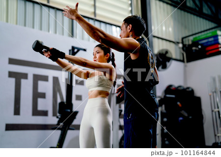 Asian sport woman hold weight to exercise with support by the trainer in fitness gym. Asian sport woman hold weight to exercise with support by the trainer in fitness gym. 101164844