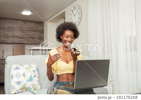 African American black woman records voice message, phone and laptop, working from home indoor. Communication with friends online, internet. Smiling 101170208