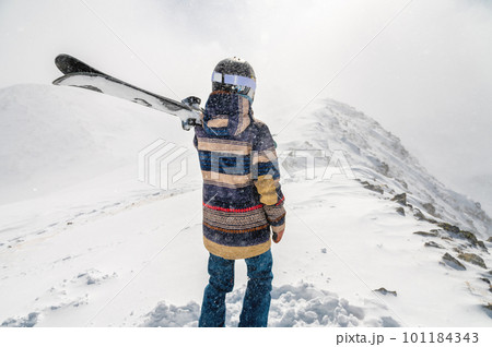 The skier stands on the top of the mountain and enjoys the view of the beautiful winter mountains on a cloudy day. Woman in ski suit holding skis on her shoulder 101184343