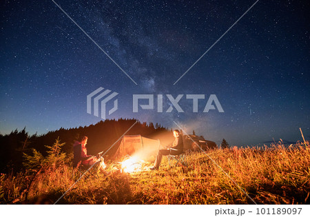 Night camping in the mountains under starry sky full of stars. Travelling couple having a rest at campfire, tourist tent and off-road car in the evening on mountain lawn and stargazing together. 101189097