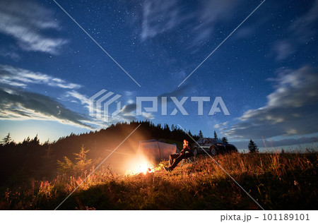 Camping in mountains under evening cloudy sky. Young man having a rest next to campfire, near tourist tent and off-road vehicle on hill. Travelling man sitting and stargazing at night. Camping in mountains under evening cloudy sky. Young man having a rest next to campfire, near tourist tent and off-road vehicle on hill. Travelling man sitting and stargazing at night. 101189101