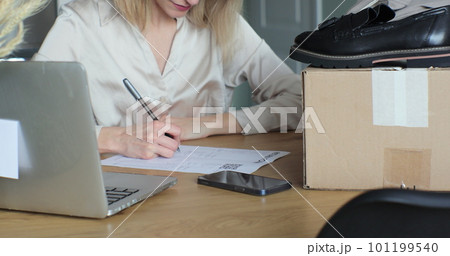 Young woman filling out a paper form, wanting to return shoes. Female writes a return order and sits at a table with a laptop at home. 101199540