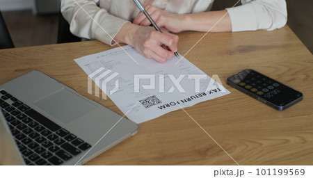 Young woman is writing tax return form and looking at laptop at table in home room. Close up woman fills out paper document and counts on the phone screen. Young woman is writing tax return form and looking at laptop at table in home room. Close up woman fills out paper document and counts on the phone screen. 101199569