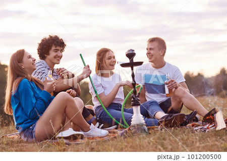 Two couple have joy together while have picnic outdoor, smoke hookah, drinks energetic beverages, being in high spirit, tell each other funny life stories. Outdoor shot. Relationship concept 101200500