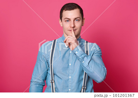 Mysterious handsome model puts his forefinger in front of mouth, showing shh sign, keeps in silent, looks curious. Brunette magnetic European boy stands in studio, looking directly at camera. Mysterious handsome model puts his forefinger in front of mouth, showing shh sign, keeps in silent, looks curious. Brunette magnetic European boy stands in studio, looking directly at camera. 101201367