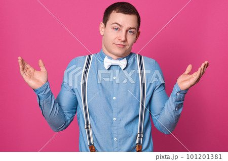 Close up portrait of careless guy spreading hands in helpless gesture, young man wearing blue shirt, suspenders and white bowtie shrugging shoulders, posing isolated over pink studio background. 101201381