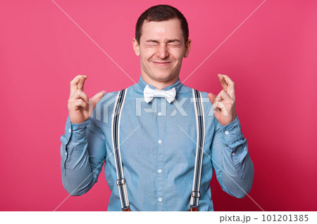 Positive young dark haired male with charming smile, has short hairstyle, crosses fingers, wishes good luck, hopes for something, screws his eyes isolated over rosy background. Body language concept. Positive young dark haired male with charming smile, has short hairstyle, crosses fingers, wishes good luck, hopes for something, screws his eyes isolated over rosy background. Body language concept. 101201385