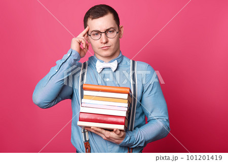 Thoughtful confused busy young model holds bunch of books in one hand, touches temple with forefinger, looks aside, wearing casual clothes, isolated over pink background in studio. Study concept. 101201419