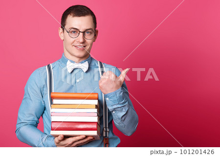 Positive sincere model looks directly at camera holding huge stack of books with one hand, showing direction with thumb, looks motivated, wearing smart casual clothes. Copy space for advertisement. Positive sincere model looks directly at camera holding huge stack of books with one hand, showing direction with thumb, looks motivated, wearing smart casual clothes. Copy space for advertisement. 101201426