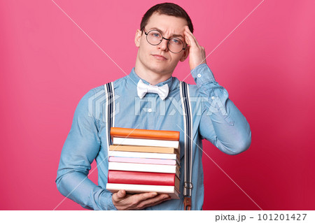 Image fo young student man holding stack of books and having headache, keeps hand on head, model poses on rose studio background, looks directly at camera. Copy space for your text. Education concept. Image fo young student man holding stack of books and having headache, keeps hand on head, model poses on rose studio background, looks directly at camera. Copy space for your text. Education concept. 101201427